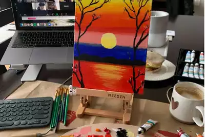 Creative teambuilding activity setup featuring a vibrant painting of a sunset with a laptop showing a virtual meeting, art supplies, and a cup of coffee. Ideal for HR professionals planning engaging remote or in-person team events.