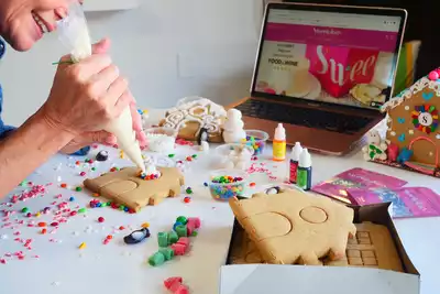 A person decorates a gingerbread house with icing and colorful candies, surrounded by various decorating supplies. A laptop displaying a sweets-themed website is visible in the background. Ideal for HR professionals seeking engaging team-building activities.