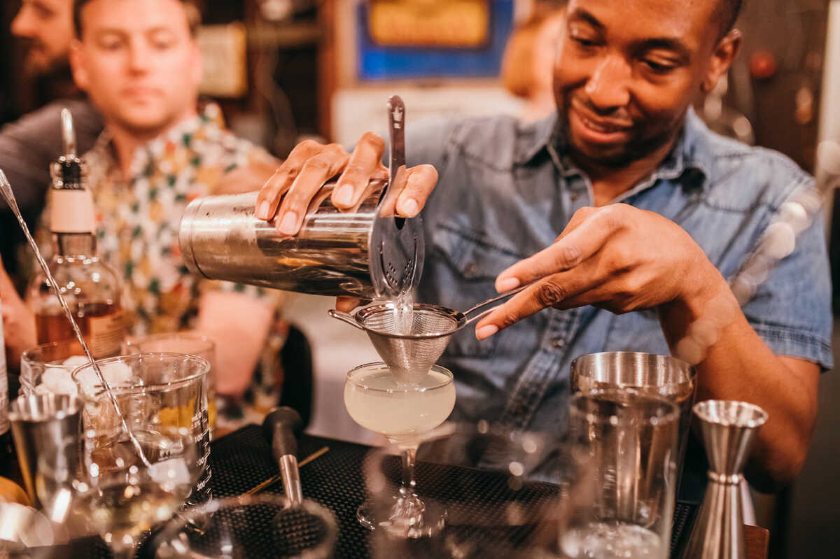 A person skillfully pours a cocktail through a strainer during a mixology workshop, surrounded by glasses and bottles. A small group observes, creating an interactive and engaging team-building experience.