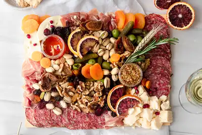 Charcuterie board featuring an assortment of cured meats, cheeses, nuts, dried fruits, fresh olives, and sliced citrus, elegantly arranged with herbs. Accompanied by wine and crackers, highlighting a gourmet experience perfect for team-building events and corporate gatherings.