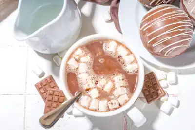 A cozy setup featuring a mug of hot chocolate topped with marshmallows, a plate of chocolate bomb treats, a jug of milk, and chocolate squares scattered around. Perfect for team-building events focused on creating a warm and inviting atmosphere for HR professionals and employees.