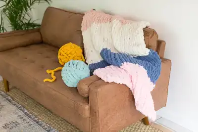 A cozy brown sofa with a chunky knit blanket in pink, white, and blue draped over its side. Large yarn balls in yellow and light blue rest on the seat, creating a welcoming atmosphere perfect for teambuilding activities focused on creativity and relaxation.