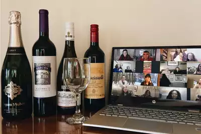 A selection of wine bottles and a wine glass displayed on a table next to a laptop screen showing a virtual meeting with smiling participants. Perfect setup for a virtual team-building wine tasting event.