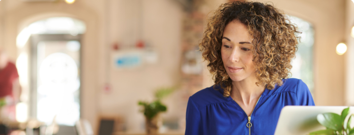 A professional in a blue blouse working on a laptop in a bright, casual workspace, surrounded by paperwork and a coffee cup, representing an HR professional planning team-building activities.