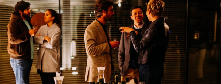 A group of people engaged in conversation and enjoying drinks at an indoor event, featuring a warmly lit space designed for teambuilding activities. A table in the foreground displays bottles and decorations, creating an inviting atmosphere for networking and collaboration.