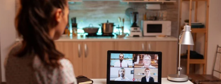 A woman participates in a virtual team-building event using a laptop in a kitchen setting. The screen displays four colleagues in an online meeting, highlighting a professional video conference environment. A notebook and lamp are on the desk, suggesting a focus on remote work and communication for HR professionals.