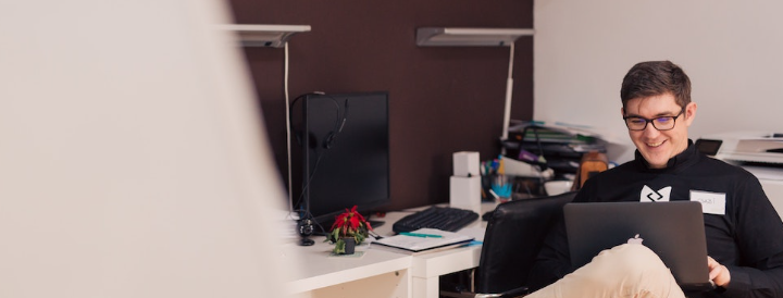 A person sitting at an office desk, smiling while working on a laptop. Ideal setting for teambuilding activities featured on a website aimed at HR professionals.