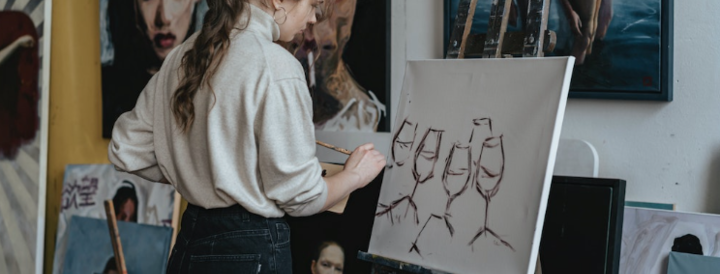 A woman in a cozy sweater and jeans stands at an easel, painting a canvas with an artistic depiction of wine glasses. The setting is an art studio filled with various paintings and creative works. This image illustrates a creative team-building activity offered for HR professionals seeking engaging events.