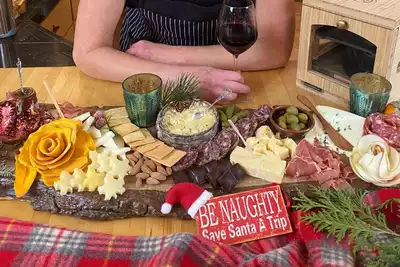 A beautifully arranged charcuterie board featuring a variety of cheeses, meats, nuts, and fruits, ideal for a team-building event. A person is seated with a glass of red wine in the background, creating an inviting and festive atmosphere. A cheeky sign reading Be Naughty, Save Santa a Trip adds humor to the setting. Perfect for HR professionals looking to book engaging team-building activities.